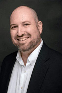 Smiling man in a black suit and white shirt against a dark background.